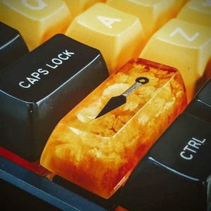 May include: A custom keyboard keycap with a golden yellow and orange resin design. The keycap features a black silhouette of a throwing knife with a ring on the handle. The keycap is on a black keyboard with yellow keys.