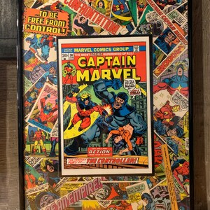 May include: A framed comic book cover featuring Captain Marvel in a red and blue costume, fighting a villain in a black and blue costume. The cover is surrounded by other comic book covers, creating a collage effect. The text on the cover reads "Captain Marvel" and "The Controller!"