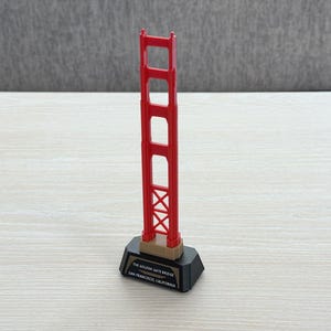 Könnte beinhalten: Ein Miniatur-Souvenir der Golden Gate Bridge in Rot mit schwarzem Sockel. Auf dem Sockel steht "THE GOLDEN GATE BRIDGE SAN FRANCISCO, CALIFORNIA". Die Brücke ist eine detaillierte Nachbildung.