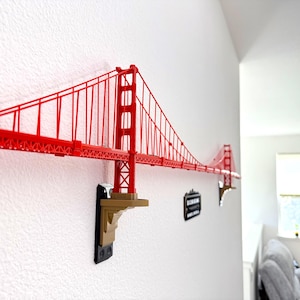 Könnte beinhalten: Rote Wanddekoration des Golden Gate Bridge. Die Brücke ist an einer weißen Wand mit dekorativen Halterungen befestigt. Die Brücke ist eine detaillierte Nachbildung des Wahrzeichens.