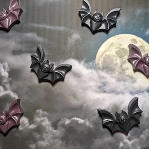 May include: Six purple and black bat-shaped soap bars against a backdrop of a cloudy night sky with a full moon.