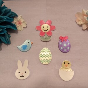 May include: A set of six Easter-themed decorations, including a blue bird, a white bunny, a chick in an eggshell, a purple egg with a bow, a green and white striped egg, and a pink flower with a yellow smiley face.