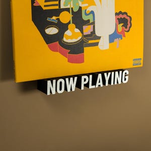 May include: A vinyl record album with a yellow cover featuring a colorful abstract design, displayed on a black shelf. The shelf has the words "NOW PLAYING" in white letters.