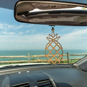 May include: A wooden pineapple car air freshener hanging from the rearview mirror of a car. The car is facing a view of the ocean and a wooden fence.