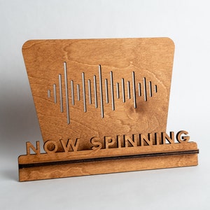 Pode incluir: Um suporte de disco de vinil de madeira com um design de onda sonora e o texto "NOW SPINNING".