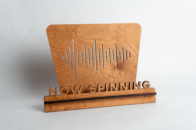 Pode incluir: Um suporte de disco de vinil de madeira com um design de onda sonora e o texto "NOW SPINNING".