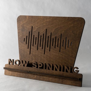 Pode incluir: Uma placa de madeira com um design de onda sonora e o texto "NOW SPINNING".