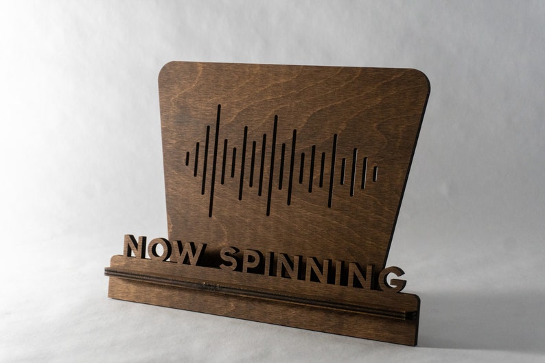 Pode incluir: Uma placa de madeira com um design de onda sonora e o texto "NOW SPINNING".