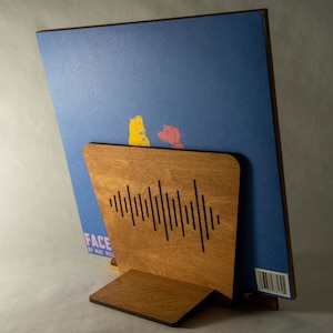 Pode incluir: Um suporte de disco de madeira com um design recortado de ondas sonoras. O suporte est&aacute; segurando um disco de vinil azul com uma ilustra&ccedil;&atilde;o amarela e vermelha na frente. O disco tem o texto "FACE" e "BY HAL WILL" na frente.