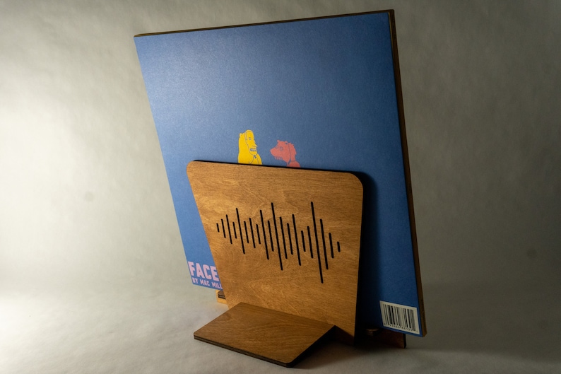 Pode incluir: Um suporte de disco de madeira com um design recortado de ondas sonoras. O suporte est&aacute; segurando um disco de vinil azul com uma ilustra&ccedil;&atilde;o amarela e vermelha na frente. O disco tem o texto "FACE" e "BY HAL WILL" na frente.