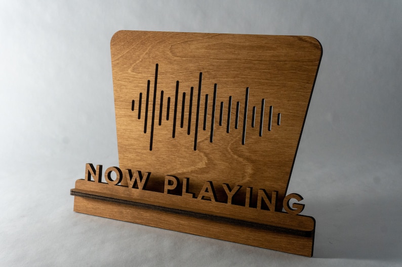 Pode incluir: Uma placa de madeira com o texto "NOW PLAYING" e um gr&aacute;fico de onda sonora. A placa &eacute; feita de madeira marrom claro e tem uma base marrom escura.
