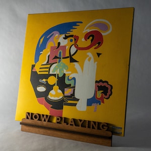Pode incluir: Um disco de vinil amarelo com uma arte abstrata colorida na capa. O disco &eacute; exibido em um suporte de madeira com as palavras "NOW PLAYING" impressas nele.