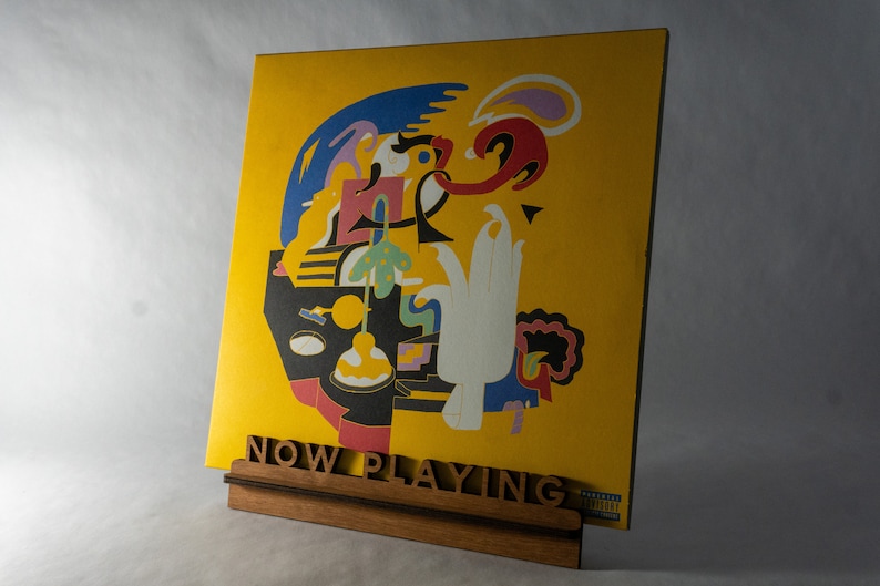 Pode incluir: Um disco de vinil amarelo com uma arte abstrata colorida na capa. O disco &eacute; exibido em um suporte de madeira com as palavras "NOW PLAYING" impressas nele.