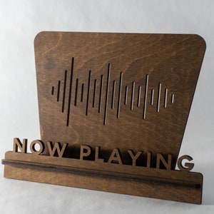 Pode incluir: Uma placa de madeira com as palavras "NOW PLAYING" e um gr&aacute;fico de onda sonora. A placa &eacute; marrom e tem um visual r&uacute;stico.