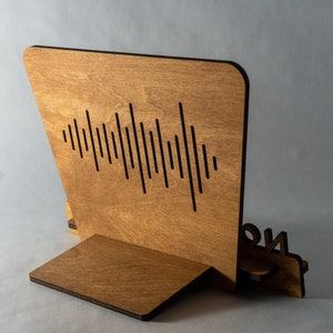 Pode incluir: Um suporte de telefone de madeira com um design de onda sonora e a palavra "ON" no lado. O suporte &eacute; feito de madeira marrom claro e tem um acabamento preto.