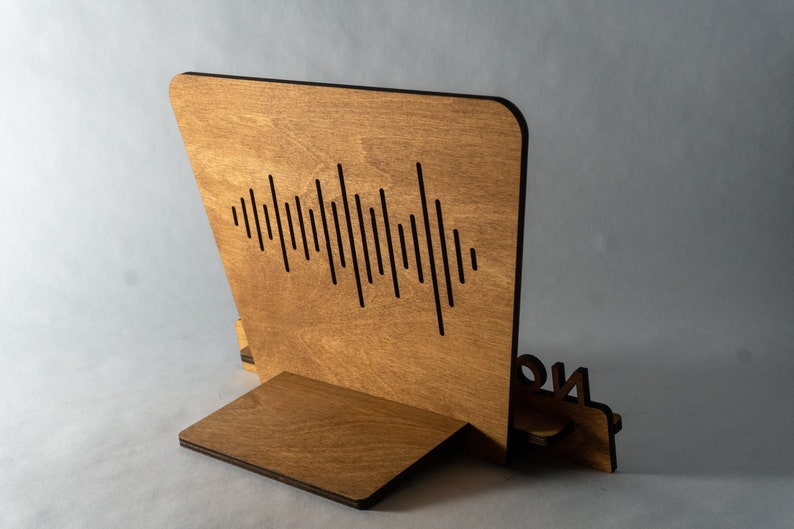 Pode incluir: Um suporte de telefone de madeira com um design de onda sonora e a palavra "ON" no lado. O suporte &eacute; feito de madeira marrom claro e tem um acabamento preto.