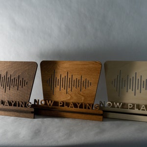 Pode incluir: Tr&ecirc;s placas de madeira com a inscri&ccedil;&atilde;o "Now Playing" e um design de onda sonora. As placas est&atilde;o em diferentes tons de madeira: marrom escuro, marrom claro e natural.
