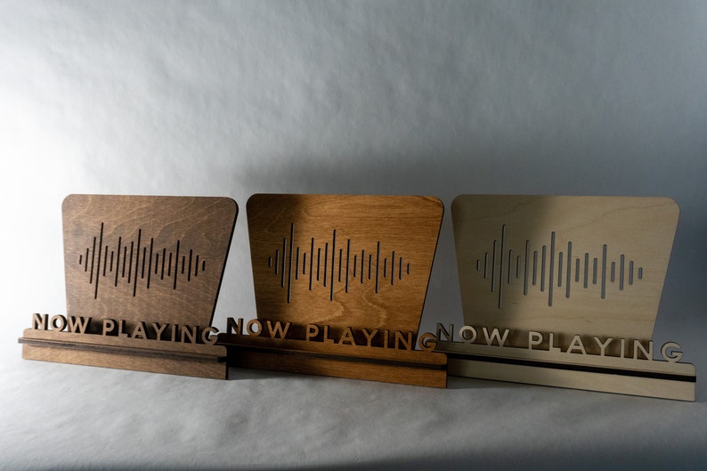 Pode incluir: Tr&ecirc;s placas de madeira com a inscri&ccedil;&atilde;o "Now Playing" e um design de onda sonora. As placas est&atilde;o em diferentes tons de madeira: marrom escuro, marrom claro e natural.