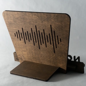 Pode incluir: Um suporte de telefone de madeira com um design de onda sonora cortado na parte de tr&aacute;s. O suporte &eacute; marrom e tem um pequeno r&oacute;tulo preto na frente que diz "ION".