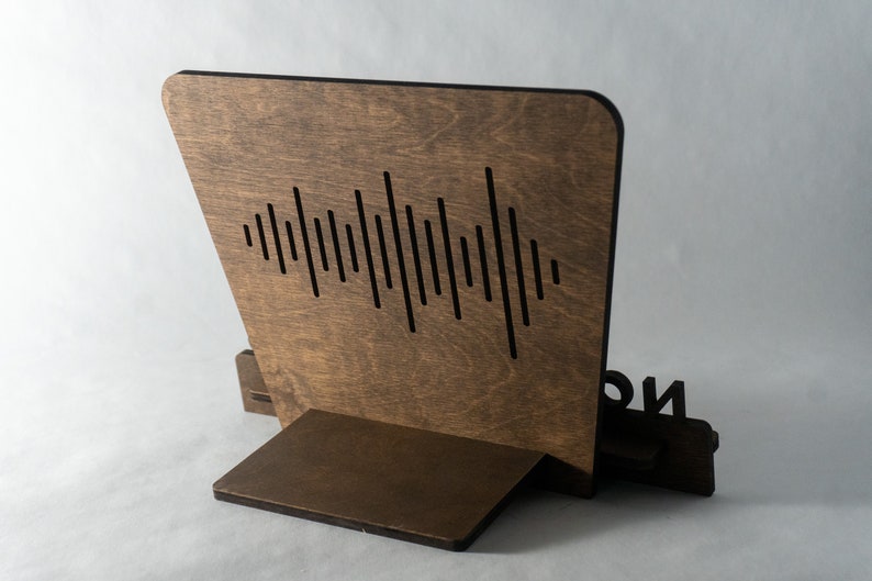 Pode incluir: Um suporte de telefone de madeira com um design de onda sonora cortado na parte de tr&aacute;s. O suporte &eacute; marrom e tem um pequeno r&oacute;tulo preto na frente que diz "ION".