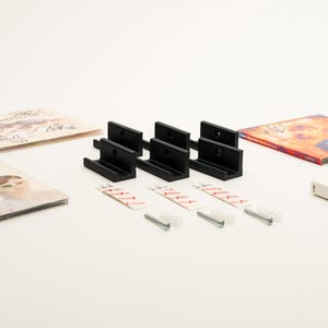 May include: Four black vinyl record wall mounts with adhesive strips and screws. The mounts are designed to hold vinyl records on a wall.