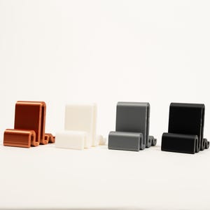 May include: Four phone stands in different colors: copper, white, gray, and black. The stands are made of a smooth, plastic-like material and have a simple, modern design.