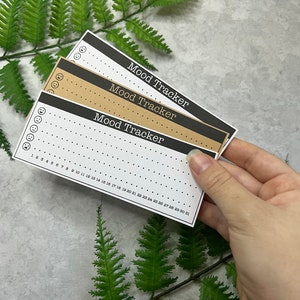 Multiple brown and white mood graph habit tracker  stickers held by a right hand on a light gray background with a green fern leaf.