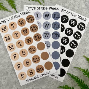 May include: Three sticker sheets with the text "Days of the Week". Each sheet features circular stickers with the days of the week abbreviated. The color schemes are neutral, blue, and black. The text "Emerald Avenue" is at the bottom of each sheet.