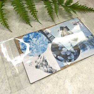 May include: A clear plastic bag filled with winter-themed paper ephemera. The ephemera includes images of snow-covered landscapes, pine trees, and a cabin. The ephemera is in shades of blue, white, and brown.