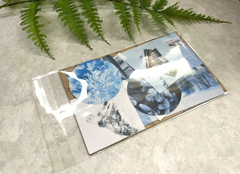 May include: A clear plastic bag filled with winter-themed paper ephemera. The ephemera includes images of snow-covered landscapes, pine trees, and a cabin. The ephemera is in shades of blue, white, and brown.
