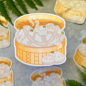 Może przedstawiać: Naklejka przedstawiająca kreskówkową ilustrację bambusowego kosza do gotowania na parze wypełnionego gotowanymi na parze pierożkami. Pierożki są beżowe z zielonymi akcentami, a para unosi się z kosza. Dodatkowe naklejki są rozrzucone.