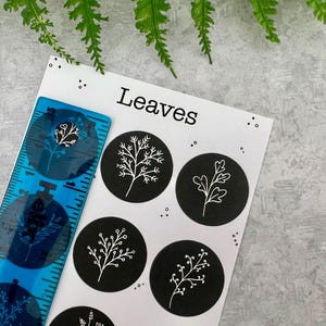 May include: A blue ruler with a floral design sits beside a white card with the word "Leaves" and black circular stickers featuring white leaf illustrations. The background is a grey surface with green leaves.