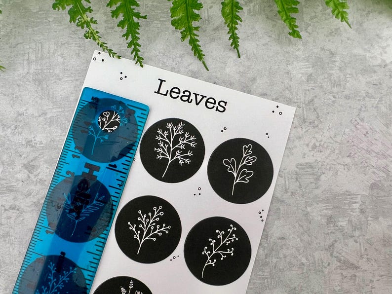 May include: A blue ruler with a floral design sits beside a white card with the word "Leaves" and black circular stickers featuring white leaf illustrations. The background is a grey surface with green leaves.