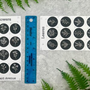 May include: Two sticker sheets with floral and leaf designs, each featuring black circles with white illustrations. The stickers are labelled "Flowers" and "Leaves" with the text "Emerald Avenue". A blue ruler is placed between the sheets.