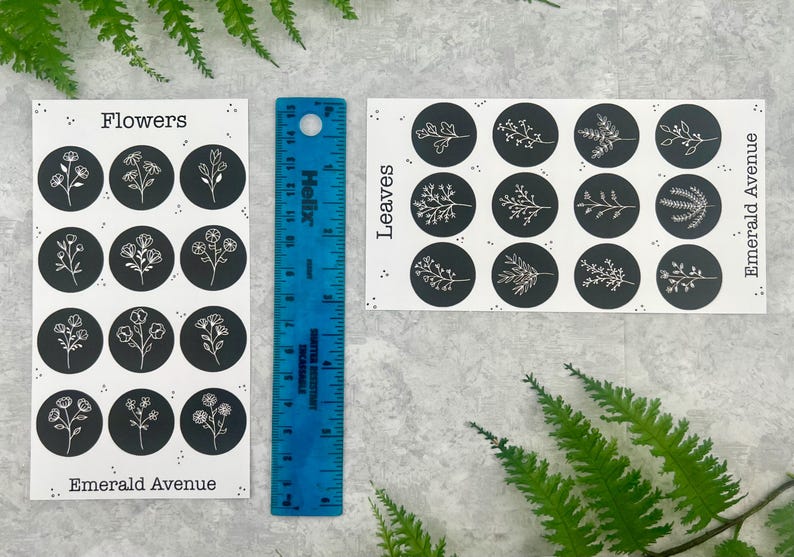 May include: Two sticker sheets with floral and leaf designs, each featuring black circles with white illustrations. The stickers are labelled "Flowers" and "Leaves" with the text "Emerald Avenue". A blue ruler is placed between the sheets.