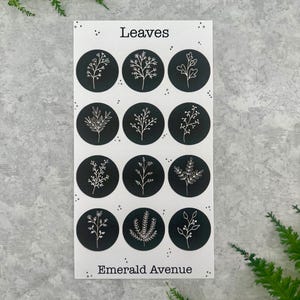 May include: A sheet of stickers with the word "Leaves" at the top and "Emerald Avenue" at the bottom. Each sticker is a black circle with a white botanical illustration. The stickers feature various leaf and plant designs.