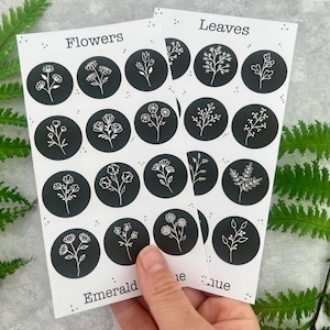May include: Two sheets of stickers featuring botanical designs. Each sticker is a black circle with a white floral or leaf illustration. One sheet is labeled "Flowers," the other "Leaves." The stickers are held by a hand, with fern leaves in the background.