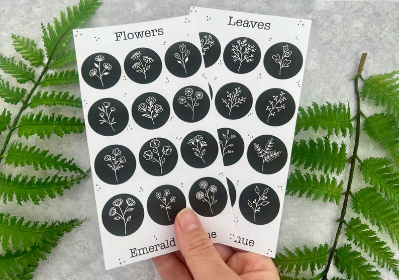 May include: Two sheets of stickers with botanical designs. Each sticker is a black circle with a white floral or leaf illustration. One sheet is labelled "Flowers," the other "Leaves." The stickers are held by a hand, with fern leaves in the background.