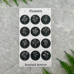 May include: A sticker sheet with the word "Flowers" at the top and "Emerald Avenue" at the bottom. The sheet features twelve black circles, each with a different white floral illustration. The background is a light grey.