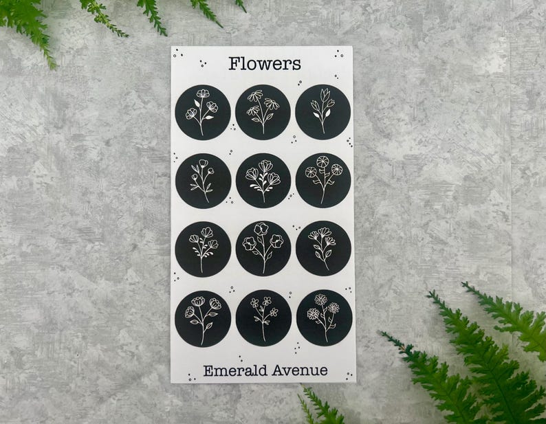 May include: A sticker sheet with the word "Flowers" at the top and "Emerald Avenue" at the bottom. The sheet features twelve black circles, each with a different white floral illustration. The background is a light grey.