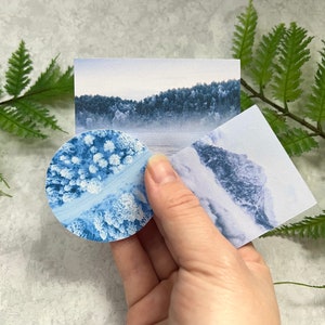 May include: Three blue and white patterned paper cards with different images of a snowy forest, a mountain range, and a forest from above. The cards are held in a hand.