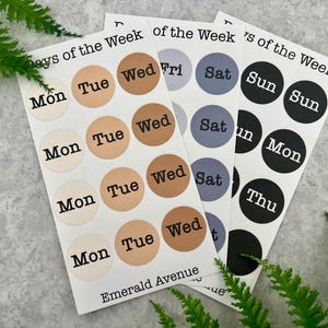 Puede incluir: Tres hojas de pegatinas con etiquetas de los días de la semana. Las pegatinas circulares muestran los días en texto negro, con fondos beige, marrón, gris y negro. El texto "Emerald Avenue" está impreso en la parte inferior de las hojas.