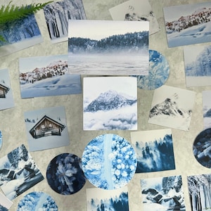 May include: A collection of winter-themed postcards and stickers featuring snowy landscapes, forests, mountains, and cabins. The images are in shades of blue and white, creating a serene and wintry atmosphere.