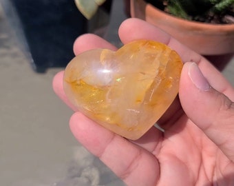 May include: A heart-shaped clear quartz crystal with golden inclusions. The crystal is polished and has a smooth, shiny surface.