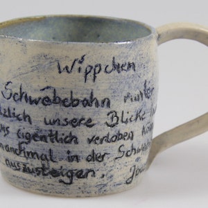 Puede incluir: Una jarra de cerámica blanca con un asa azul y texto escrito a mano en azul que dice "Wippchen, der Schwebebahn runter, holzlich unsere Blicke, manchmal verdoben hin, aussteigen. Jou"