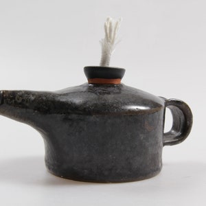 Könnte beinhalten: Eine kleine, schwarze Keramik-Öllampe mit einem weißen Docht. Die Lampe hat einen Ausguss und einen Griff.