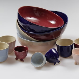 Könnte beinhalten: Ein Set aus Keramik-Schalen und -Bechern in verschiedenen Farben, darunter Blau, Rot, Rosa und Beige. Die Schalen sind übereinander gestapelt, während die Becher in einem Halbkreis um die Schalen angeordnet sind. Einige der Becher haben drei kleine Füße.