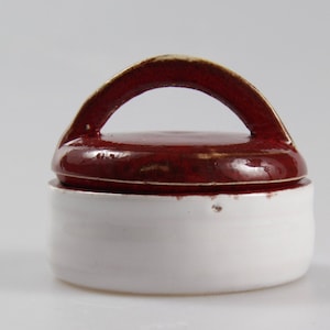 Puede incluir: Un pequeño tarro de cerámica blanca con tapa roja y asa. La tapa tiene un esmalte rojo moteado.