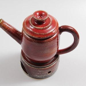 Könnte beinhalten: Eine rote Keramik-Teekanne mit Deckel und passendem Sockel. Die Teekanne hat einen runden Korpus und einen Ausguss auf einer Seite. Der Sockel hat ein Loch in der Mitte.