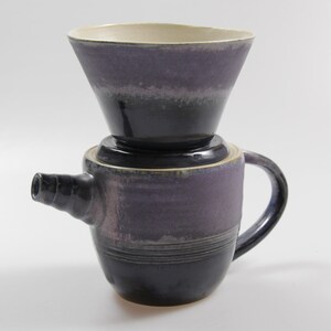 Könnte beinhalten: Eine Kaffeekanne aus Keramik mit eingebautem Filter zum Aufgießen. Die Kanne ist tiefviolett mit einem weißen Rand und einem schwarzen Henkel.